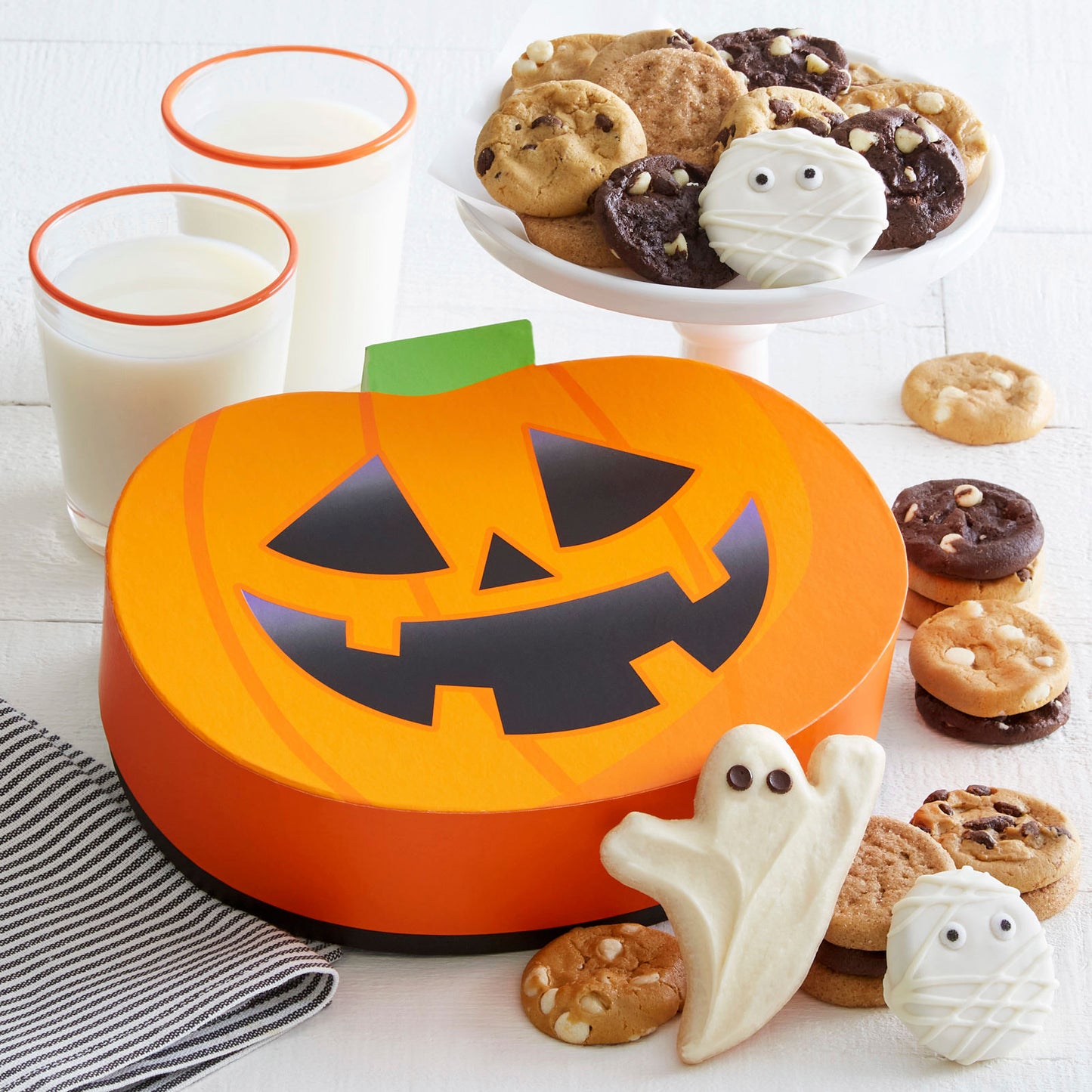 Pumpkin shaped gift box surrounded by an assortment of Nibblers®, a frosted ghost cookie, and two frosted mummy Nibbers®