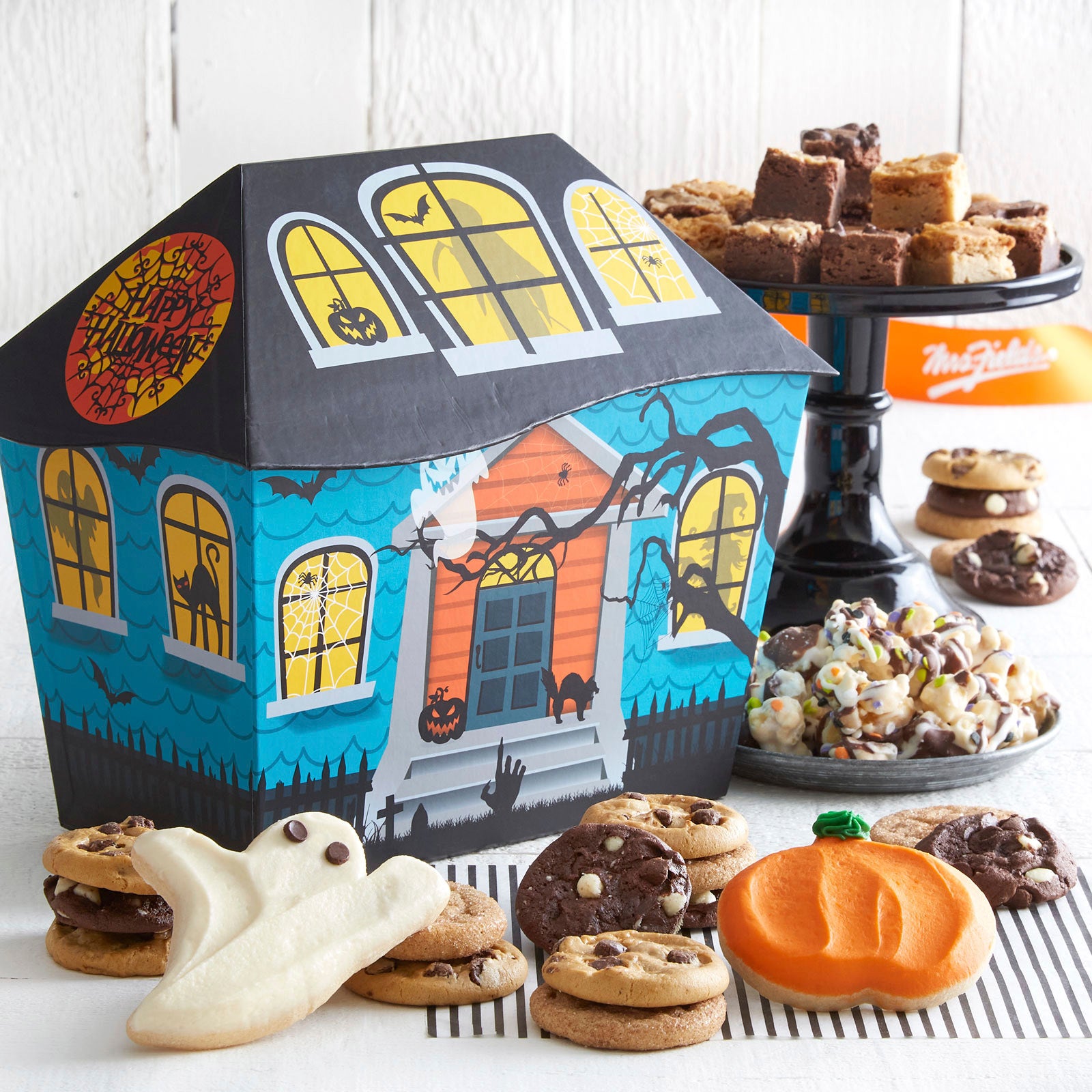 Haunted house themed gift box surrounded by an assortment of Nibblers®, brownie bites, frosted ghost and pumpkin cookie, and chocolate covered popcorn