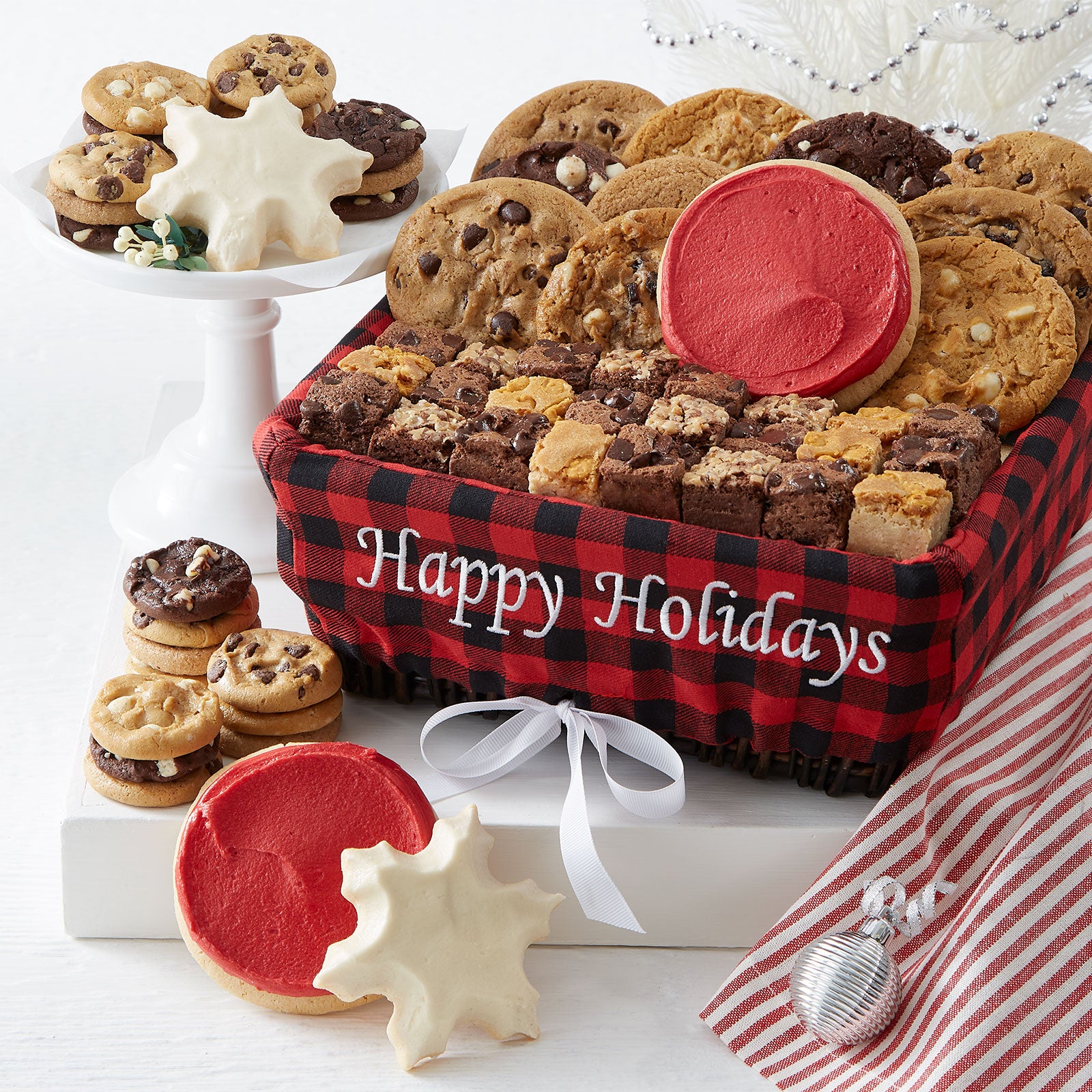 A red and black plaid Happy Holidays basket filled with an assortment of original cookies, Nibblers®, brownie bites, two frosted red round cookies, and two frosted snowflake cookies