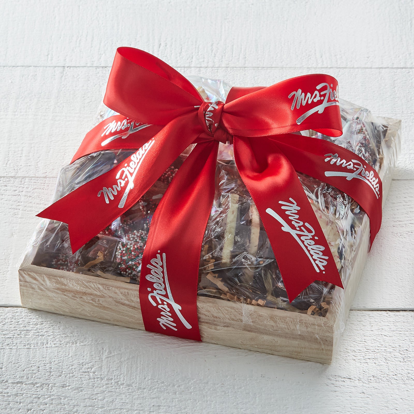 Wooden tray filled with peppermint bark and nonpareils wrapped up with a red Mrs. Fields bow.