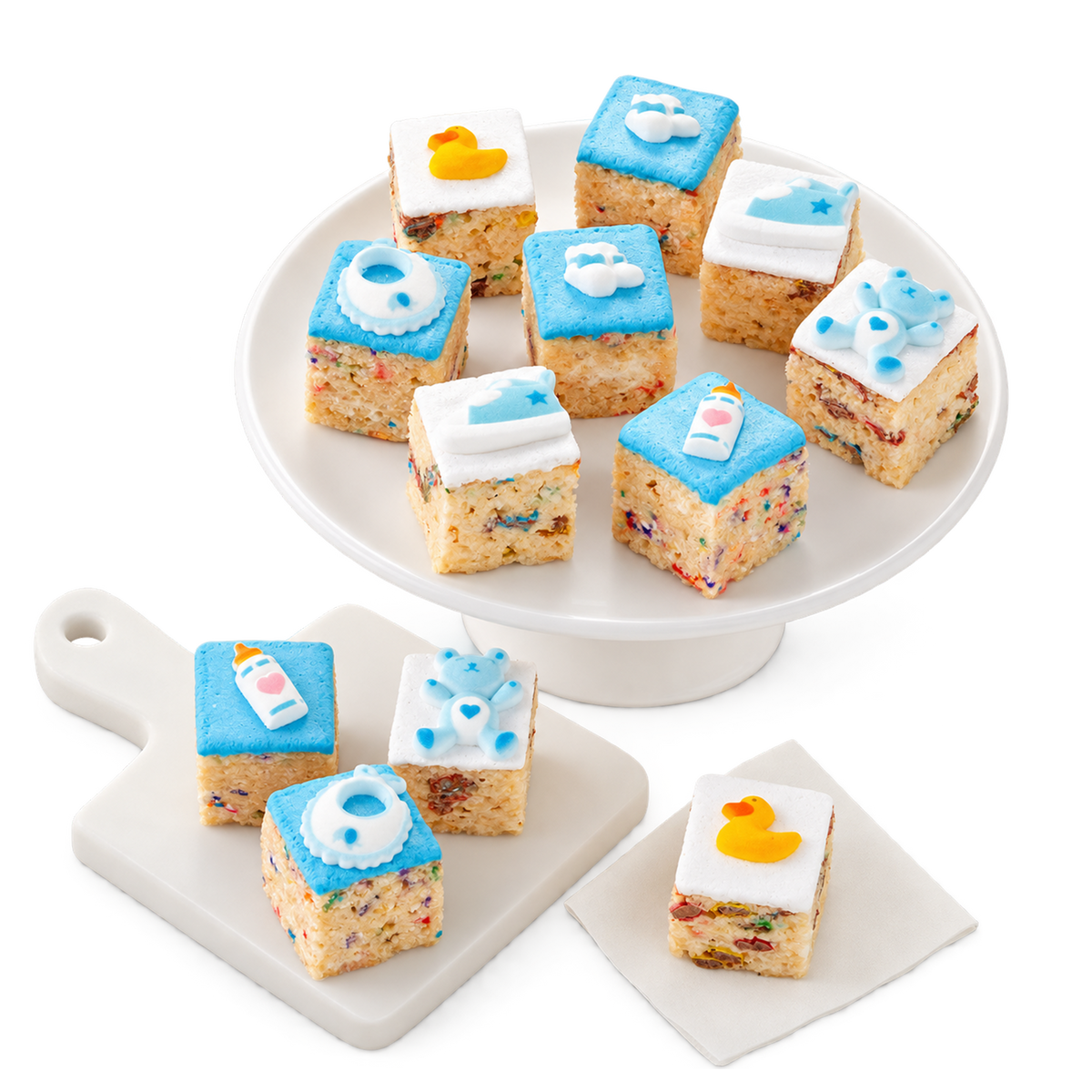 Baby-themed rice cake treats decorated to look like baby bottles, diapers, clouds, and a rubber duck, arranged on a white pedestal plate and small white serving boards against a white background.