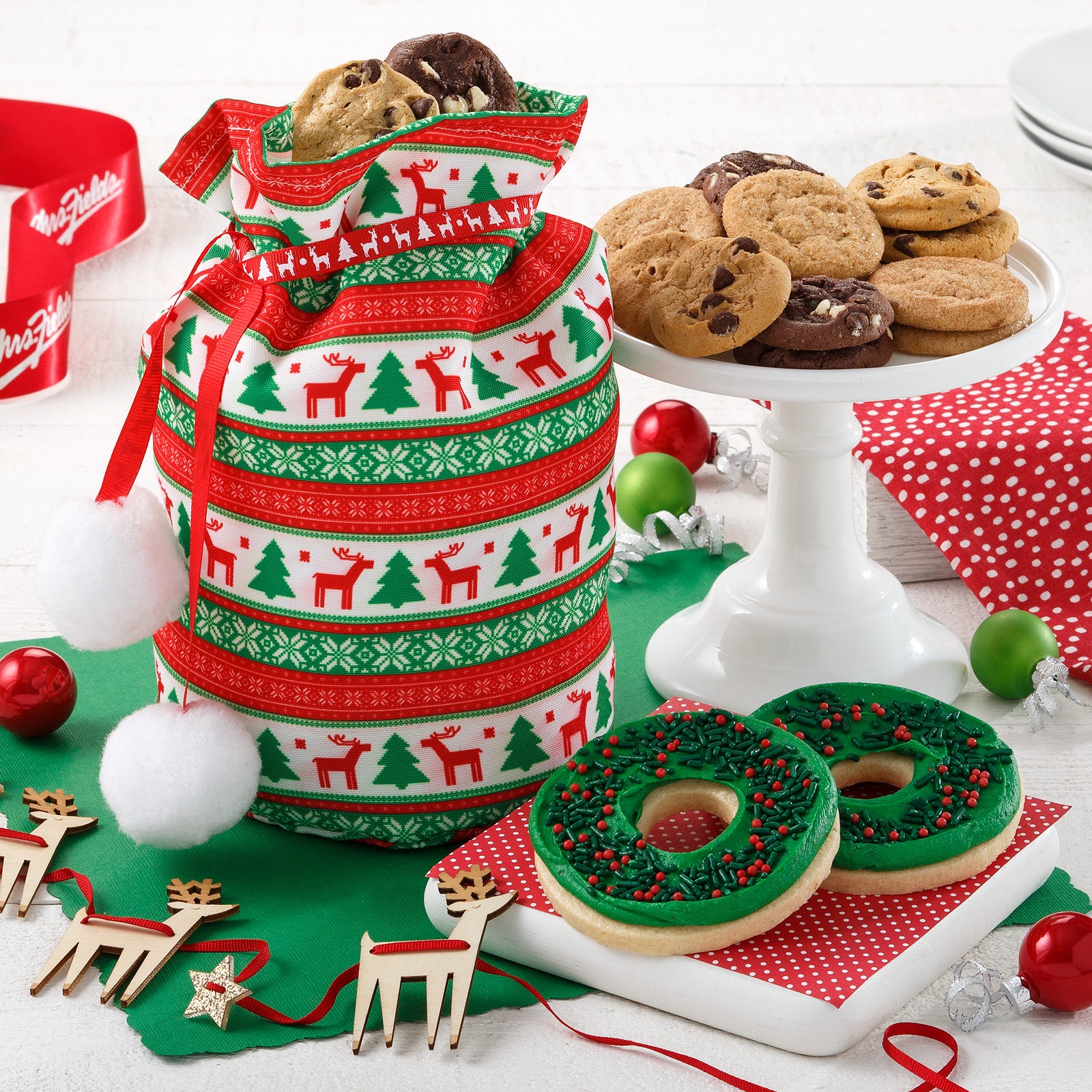 A holiday-themed tote bag with red and green decorations with an assortment of Nibblers® and two frosted wreath cookies with sprinkles