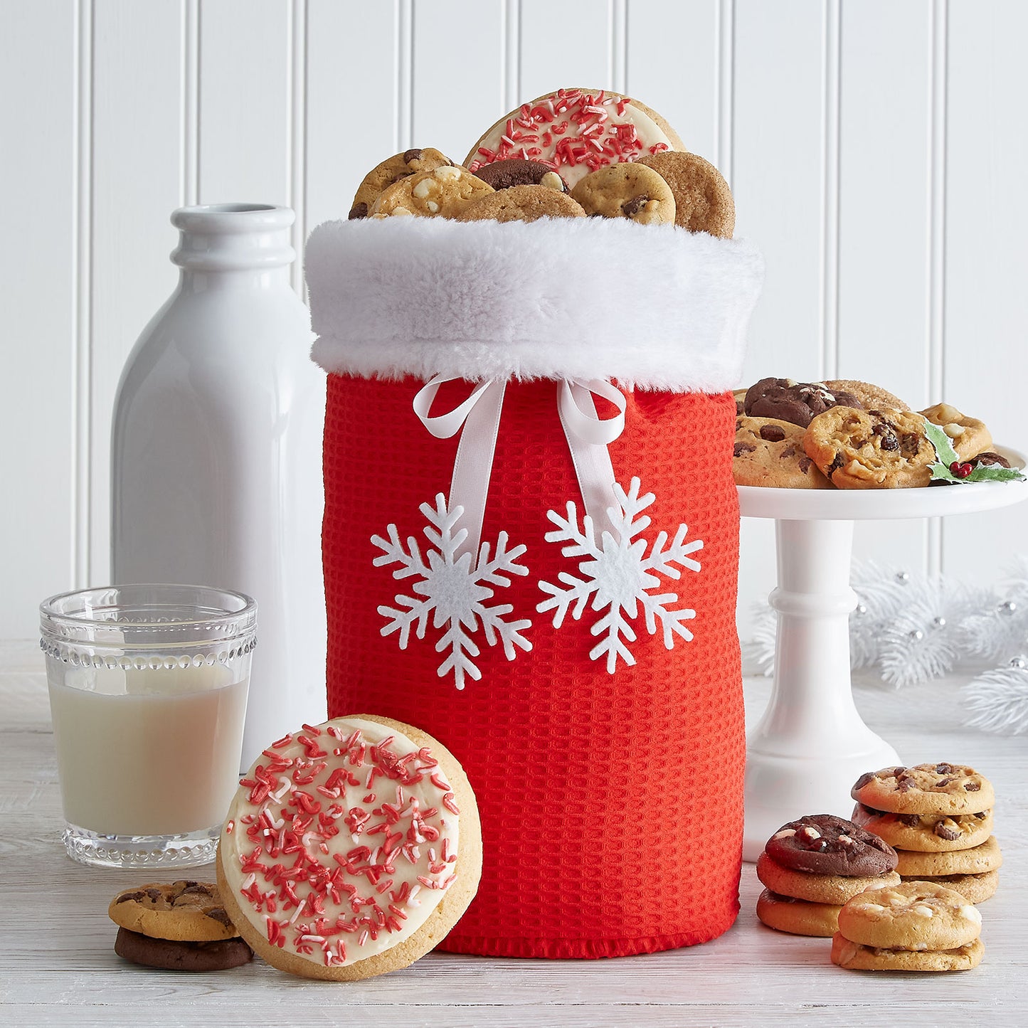 A red and white holiday tote decorated with two snowflakes and filled with an assortment of Nibblers® and two frosted peppermint round cookies.