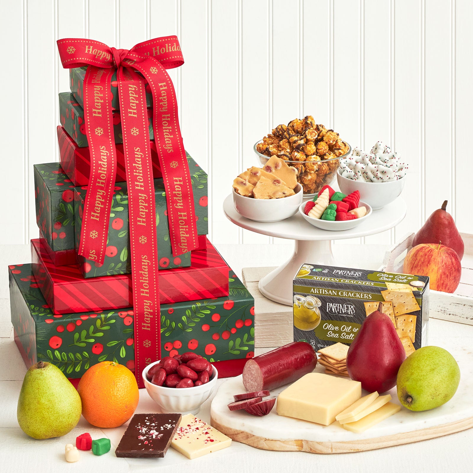 A multiple-tiered gift tower surrounded by an assortment of fruit, popcorn, frosted pretzels, peanut brittle, candy, crackers, cheese, sausage, and peppermint bark.