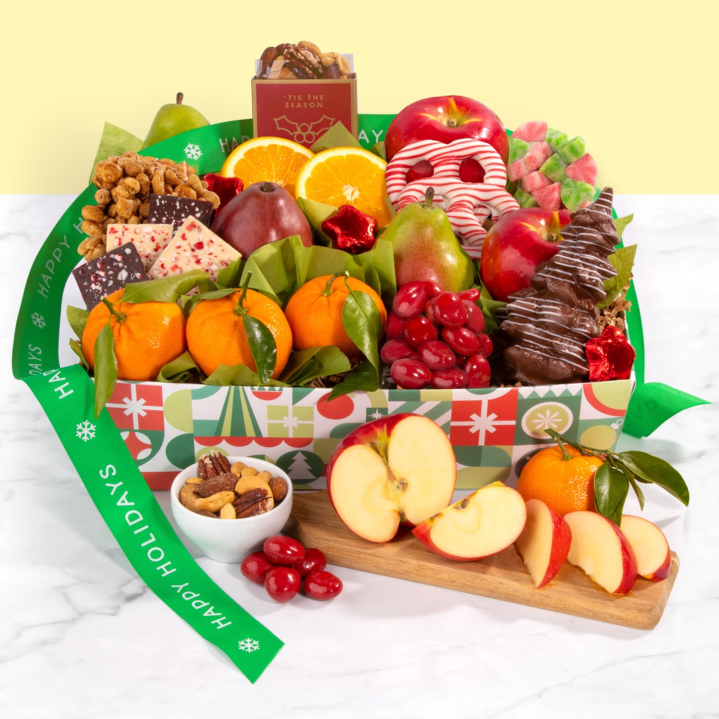 A decorated holiday crate filled with an assortment of fruit, nuts, popcorn, chocolate-covered pretzels, candy, and chocolate with a green "Happy Holidays" ribbon.