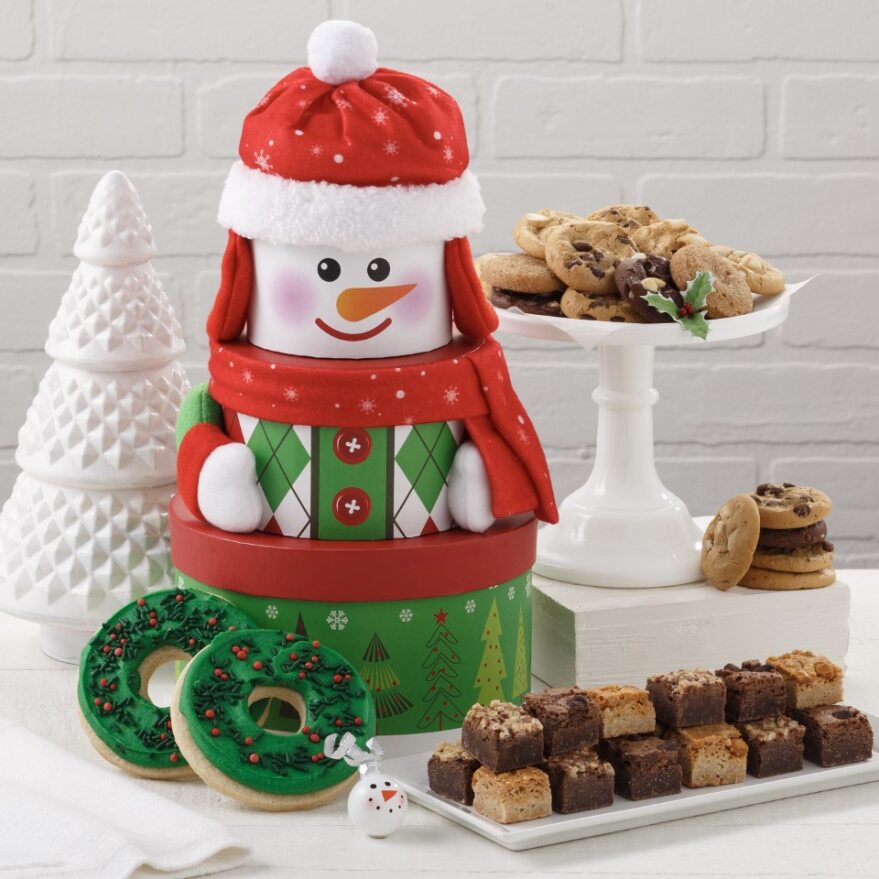 A snowman three-tier tower surrounded by an assortment of Nibblers®, brownie bites, and two frosted wreath cookies with sprinkles.