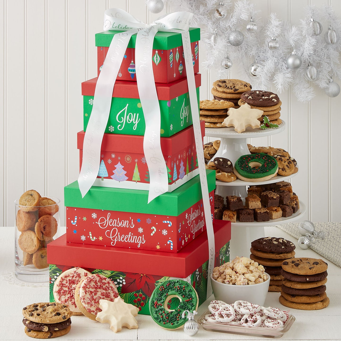 A red and green five-tier tower tied with a white Mrs. Fields ribbon and surrounded by an assortment of Nibblers®, original cookies, brownie bites, two frosted wreath cookies with sprinkles, two frosted peppermint cookies with sprinkles, two frosted snowflake cookies, popcorn, muffins, and yogurt-covered pretzels.