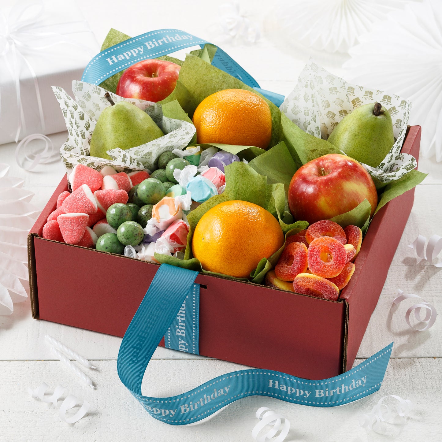 A red box filled with apples, pears, oranges and an assortment of candies with a blue happy birthday ribbon.