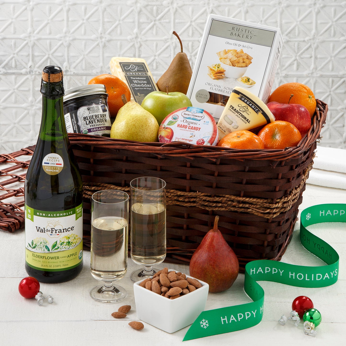 A brown gift basket filled with an assortment of fruit, nuts, cheese and a bottle of cider in front