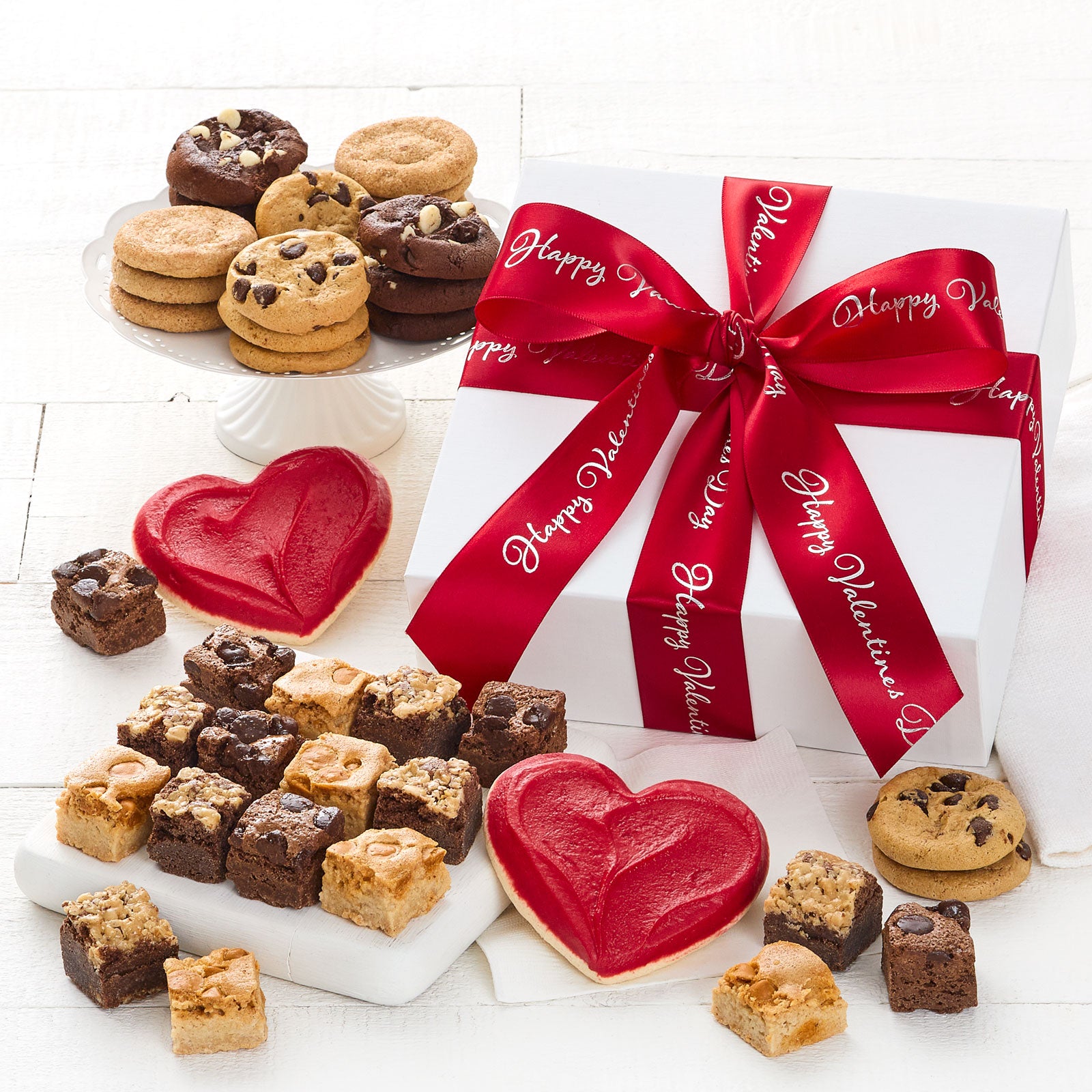 A white gift box wrapped with a Happy Valentine's Day red ribbon and surrounded with an assortment of nibblers, brownie bites, and two frosted heart cookies.