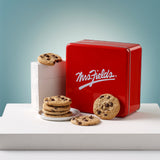 A red square metal tin with the white Mrs. Fields logo sits on a white platform, surrounded by chocolate chip cookies. A stack of cookies is on a small white plate beside the tin, and two cookies rest in front. This all sits in front of a light blue illuminated background.