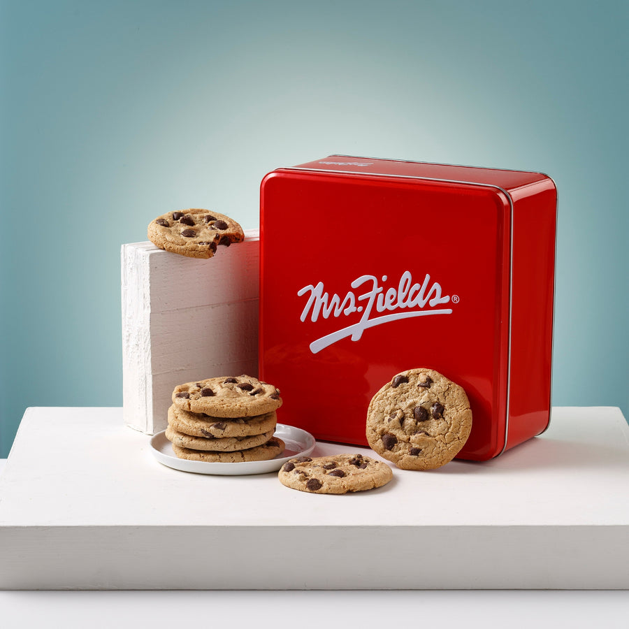 A red square metal tin with the white Mrs. Fields logo sits on a white platform, surrounded by chocolate chip cookies. A stack of cookies is on a small white plate beside the tin, and two cookies rest in front. This all sits in front of a light blue illuminated background.