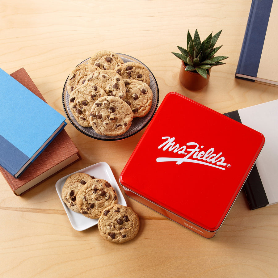 A bright red tin with the white script Mrs. Fields logo on it, styled on a light wood tabletop with chocolate chip cookies on a glass plate and a small white dish, alongside books and a potted succulent.