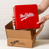 A person holds a square red tin with a white 'Mrs. Fields' logo above an open brown cardboard box printed with  the 'Mrs. Fields' logo.