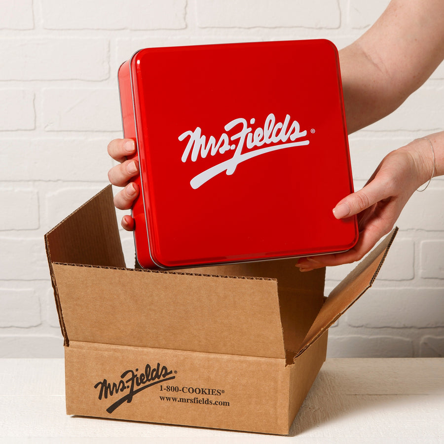 A person holds a square red tin with a white 'Mrs. Fields' logo above an open brown cardboard box printed with  the 'Mrs. Fields' logo.