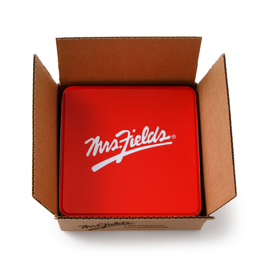 A red square metal tin with a white 'Mrs. Fields' logo, sitting inside an open brown cardboard box on a white background.