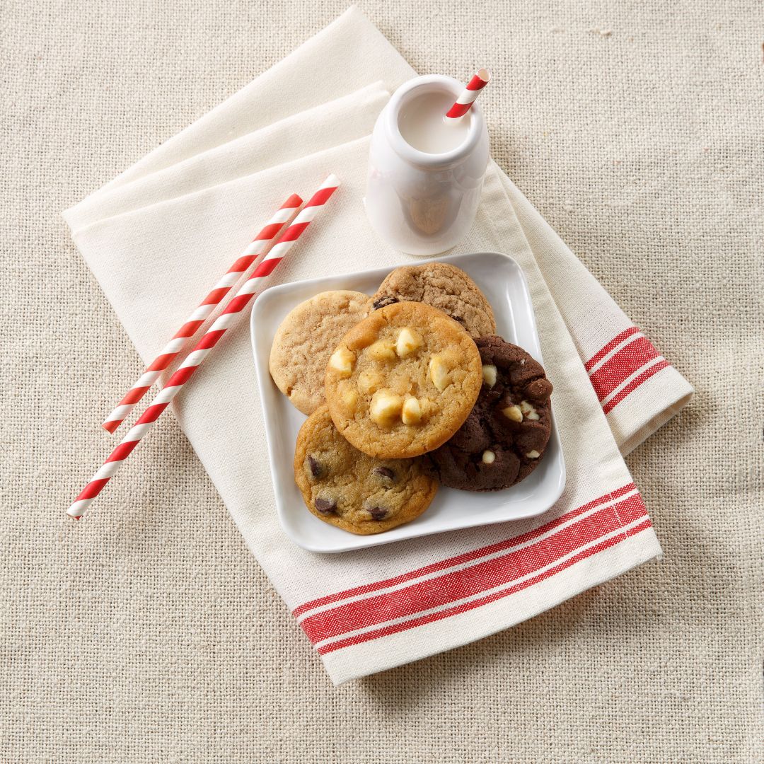 A styled flat-lay of assorted cookies on a small white plate, with a white napkin, red-striped straws, and a small white cup of milk on a beige tablecloth background.