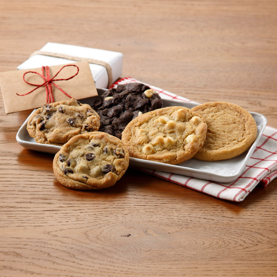 Five different cookies on a small white plate with a red and white napkin and a small envelope tied with red twine on a dark wooden tabletop surface.