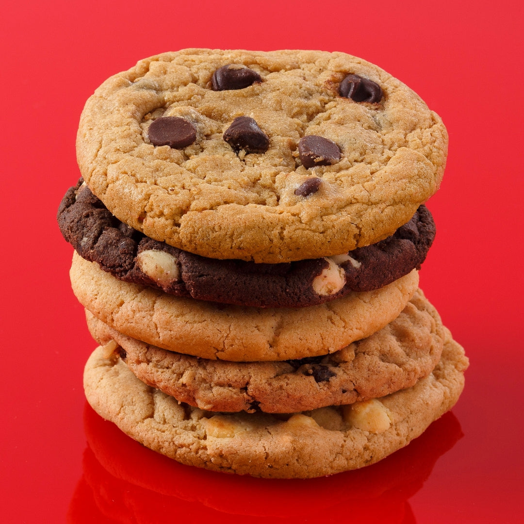 A stack of five cookies on a bright red reflective background. The top cookie is golden brown with dark chocolate chips, with four additional cookies stacked underneath, including a triple chocolate cookie, a white chocolate chip cookie, an oatmeal raisin cookie, and a white chocolate chip cookie.