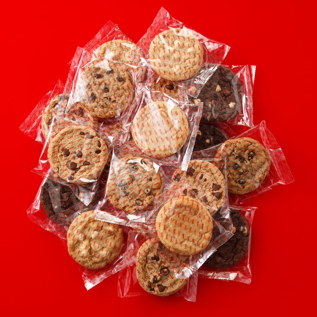 A pile of individually wrapped, assorted cookies on a solid bright red background. The clear wrapping is branded with the Mrs. Fields logo. Flavors include chocolate chip, oatmeal raisin, cinnamon sugar, triple chocolate, and white chocolate chip.