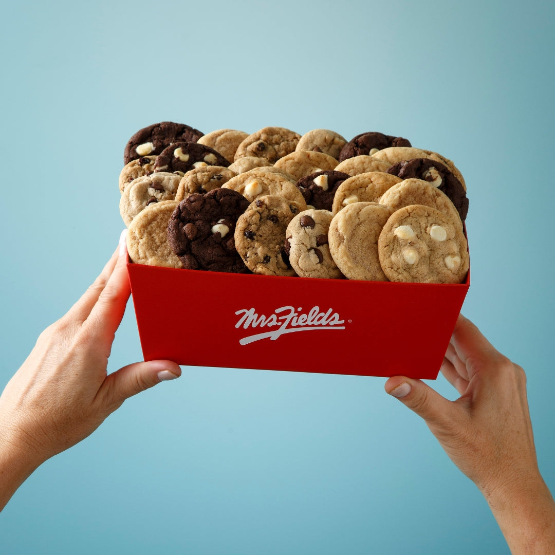 Two hands holding a red crate of assorted cookies against a light blue background. The crate has a white Mrs. Fields logo on it.