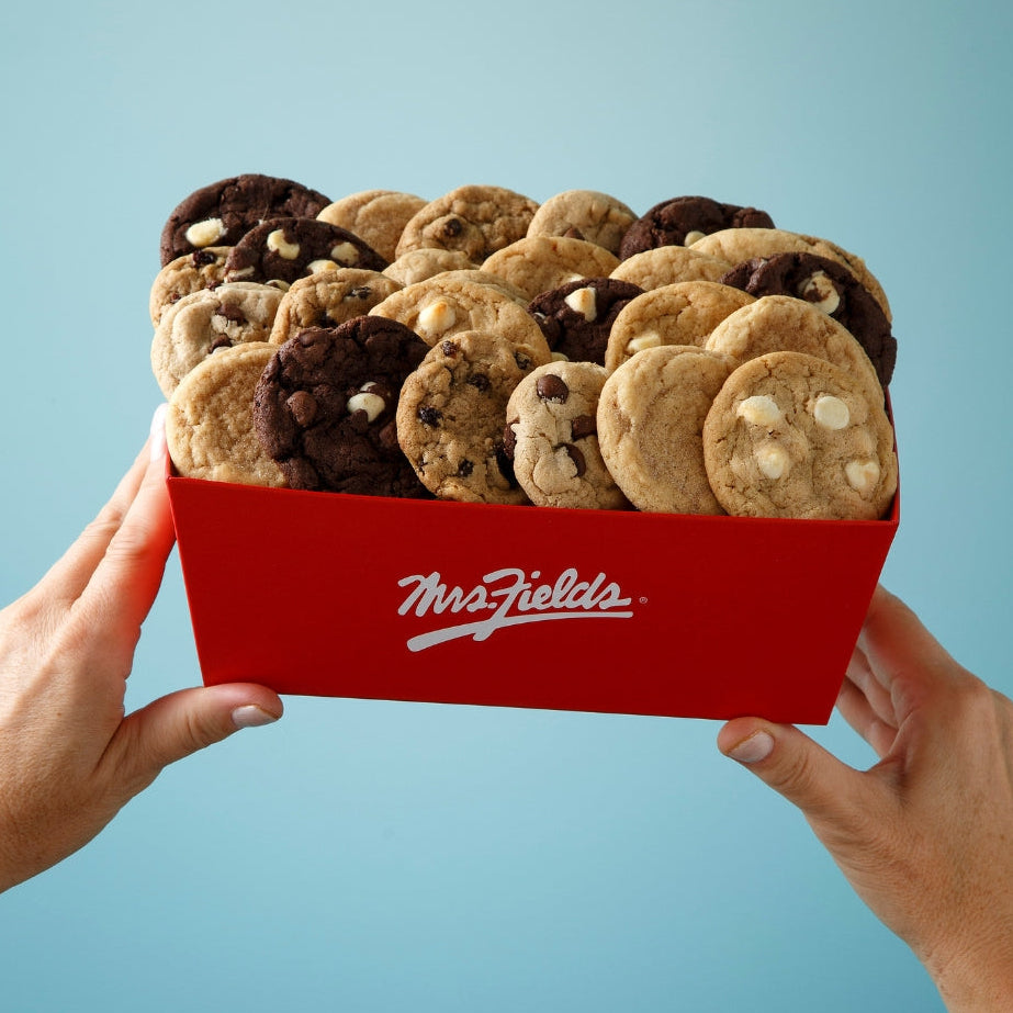 Two hands holding a red crate of assorted cookies against a light blue background. The crate has a white Mrs. Fields logo on it.