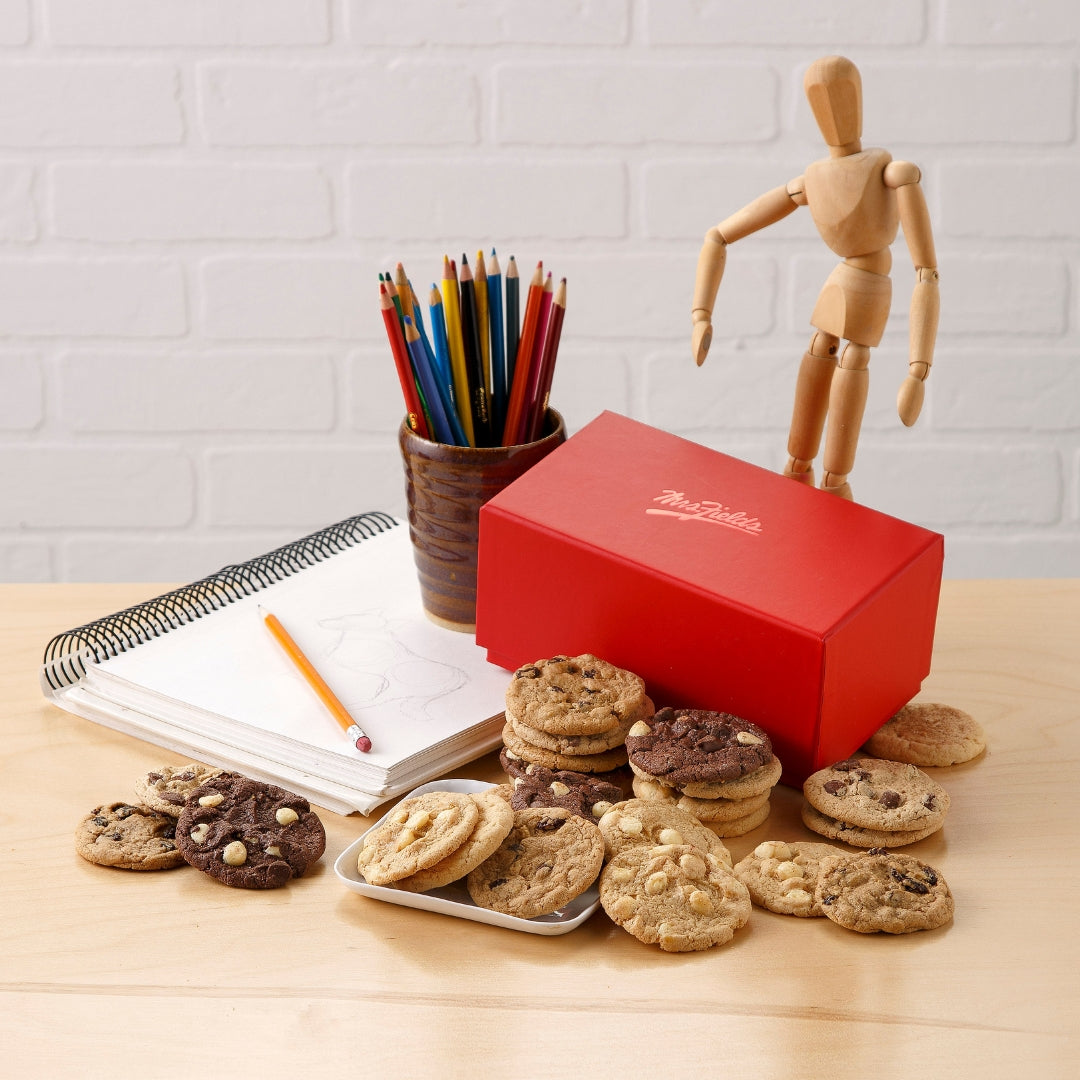 A red rectangular gift box with white script branding sits on a light wood table, surrounded by assorted chocolate chip cookies and cookie stacks. A small wooden artist mannequin stands on top of the box, next to a pencil cup with colored pencils, an open sketchbook, and a pencil.