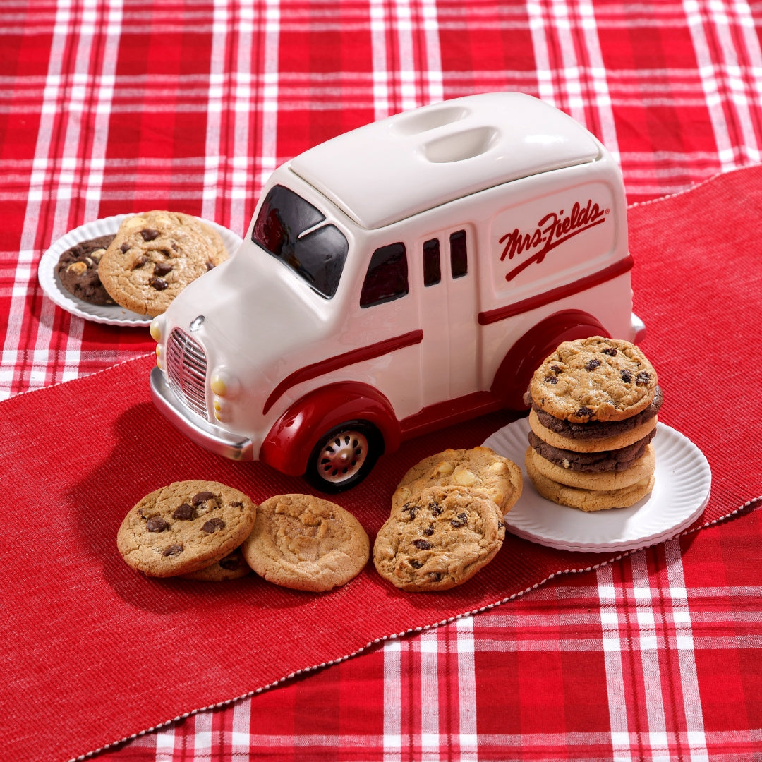 A ceramic cookie jar shaped like a vintage white and red delivery truck, labeled 'Mrs. Fields', styled with chocolate chip cookies on a red and white plaid tablecloth.