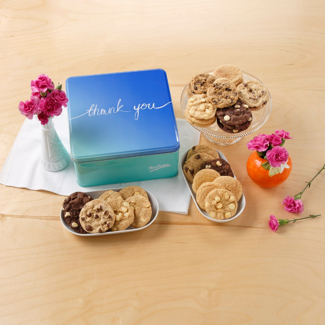 A blue gradient cookie tin with 'thank you' written on the lid, styled on a light wood table with assorted cookies in bowls and a glass plate, accented by small pink flowers.