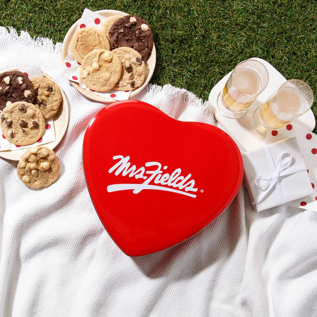 A bright red heart-shaped Mrs. Fields cookie tin sits on a white picnic blanket outdoors on green grass, surrounded by plates of cookies, two glasses of champagne, and a small gift box.