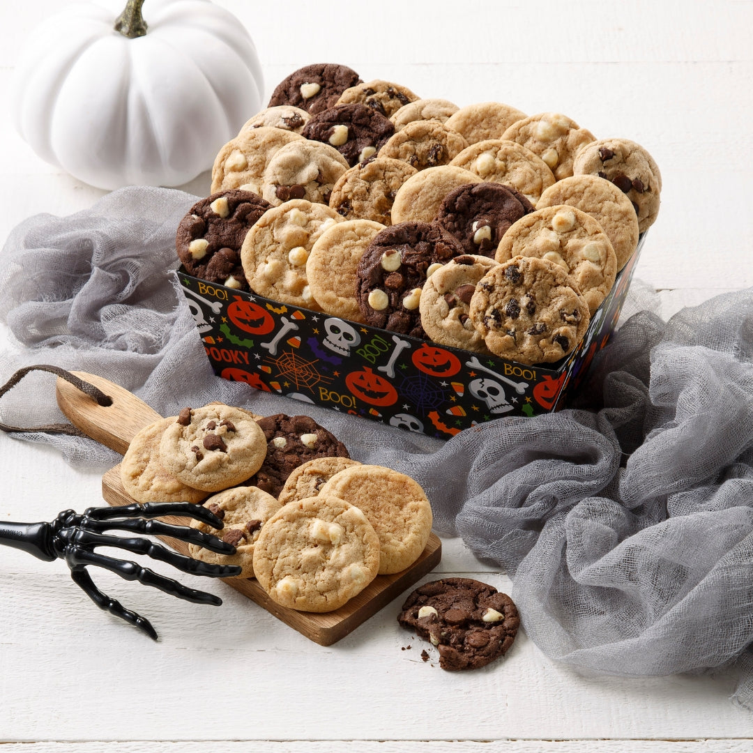 A Halloween-themed cookie assortment displayed in a decorative crate on a white wooden surface, styled with gray tulle, a white pumpkin, and a black skeleton hand prop.