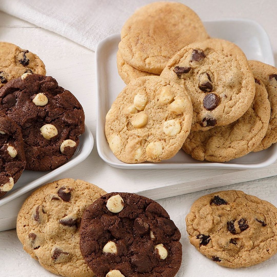 Two white square plates filled with assorted cookies on a light tabletop. One plate holds chocolate chip cookies with white chocolate chips, and the other holds dark chocolate cookies with white chocolate chips.