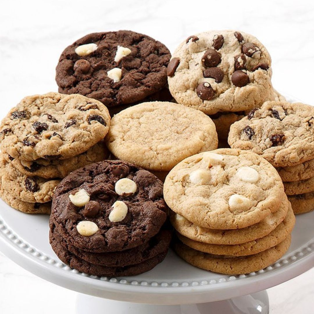 A variety of chocolate chip and white chocolate cookies arranged on a white pedestal plate against a light marble background.