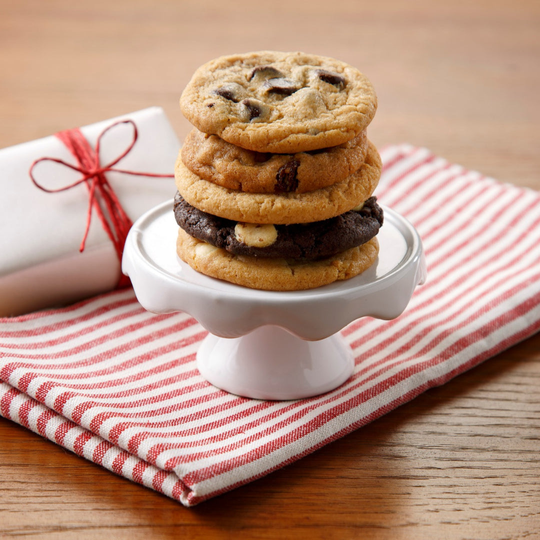A stack of five mini cookies in different flavors on a miniature cake stand atop a white and red striped cloth on a wooden surface.