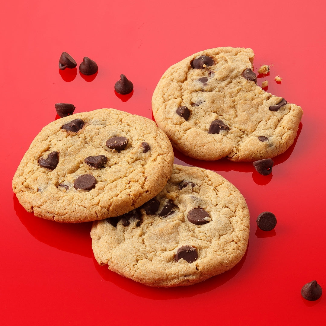 Three chocolate chip cookies are arranged on a bright red surface with chocolate chips scattered around them. One of the cookies has a bite taken out of it with a few crumbs showing.