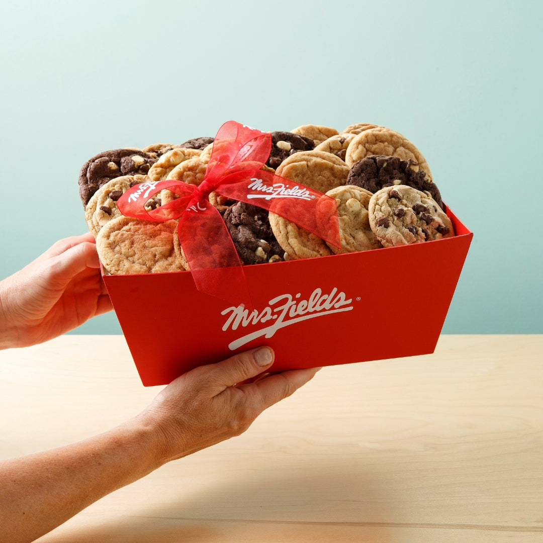 A red Mrs. Fields gift box filled with assorted chocolate chip cookies, tied with a red ribbon, held by two hands over a light wood table against a pale blue background.