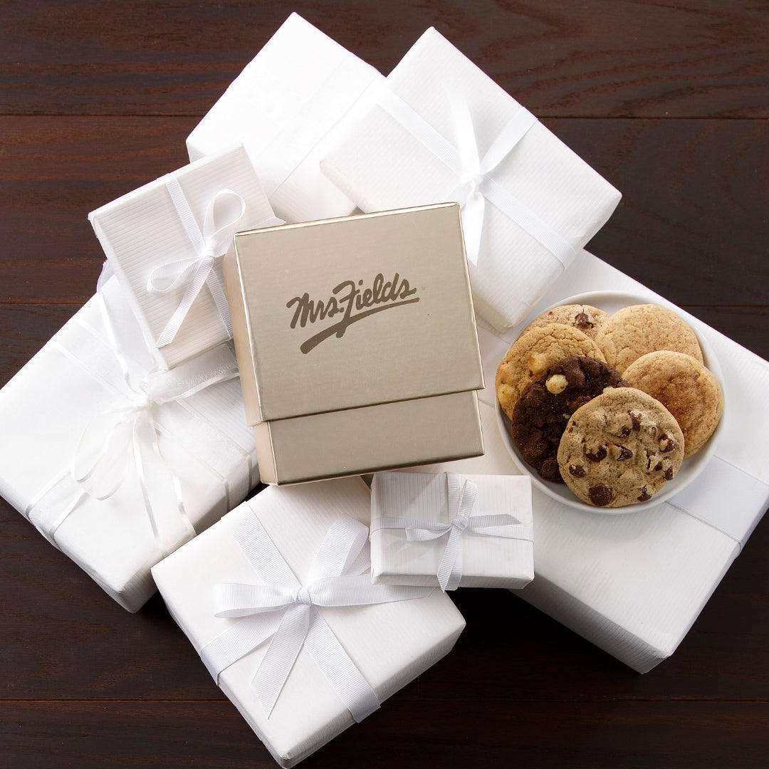 A gift set featuring a central gold square box with the 'Mrs. Fields' logo, surrounded by several wrapped gift boxes in white paper with white ribbons, and a small white plate filled with assorted cookies including chocolate chip and cinnamon sugar, all arranged on a dark wooden surface.