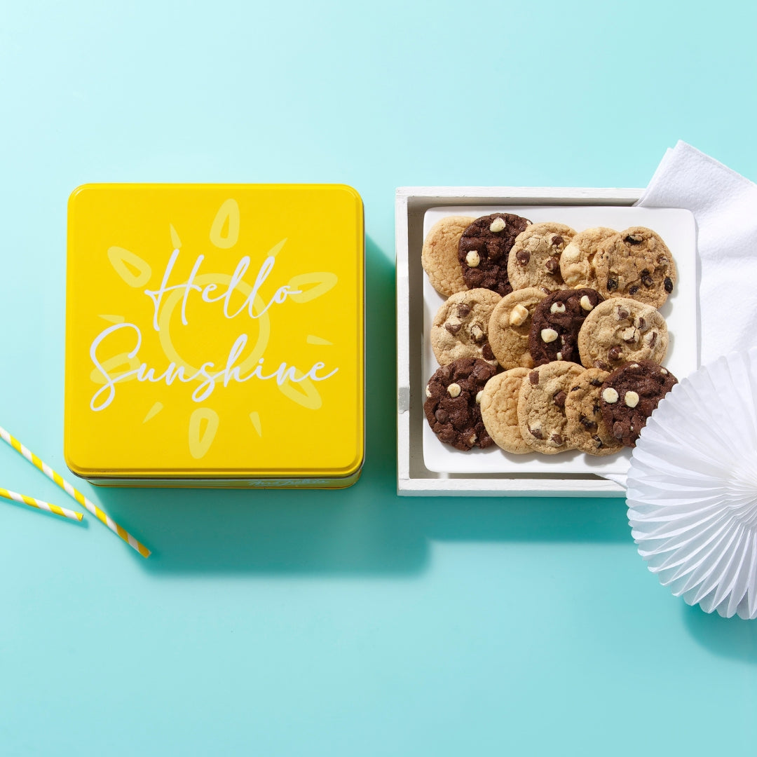 A yellow square cookie tin with 'Hello Sunshine' printed on the lid sits beside an open white tray filled with assorted chocolate chip cookies on a light blue background.