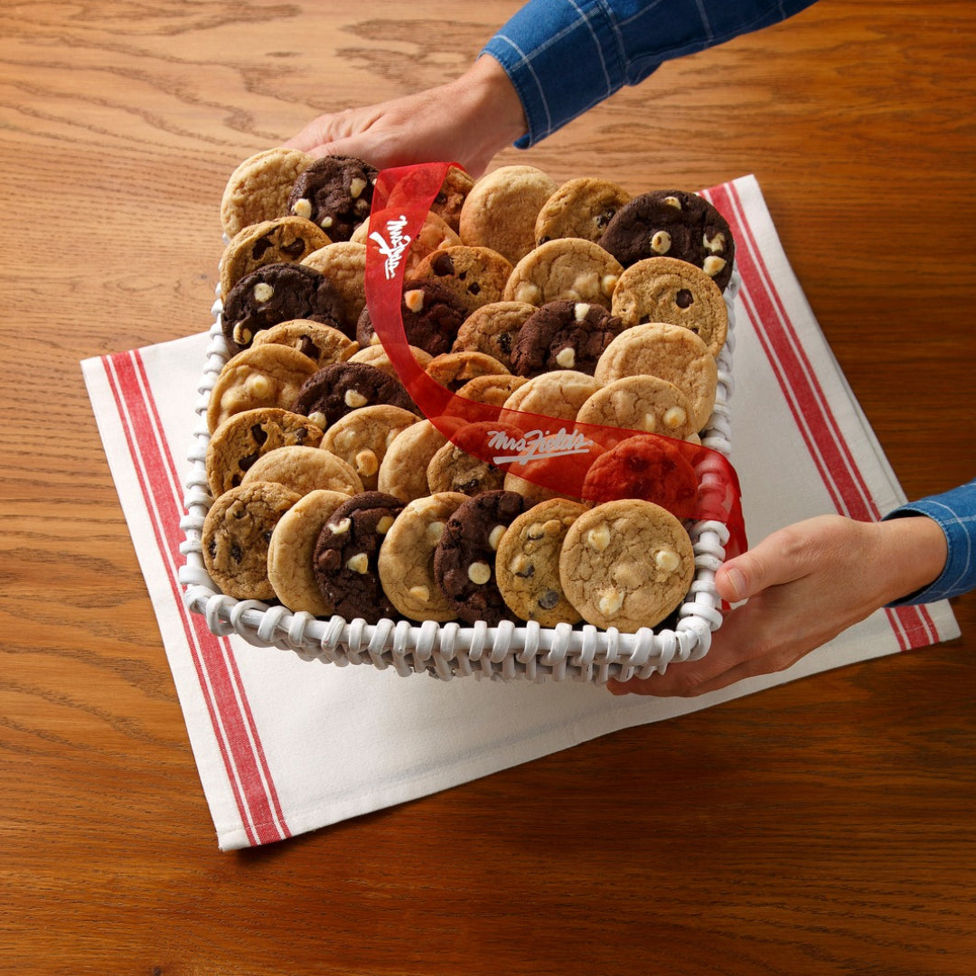 A medium sized basket filled with cookies held by a person's hands over a wooden tabletop surface with a white and red towel laid out.