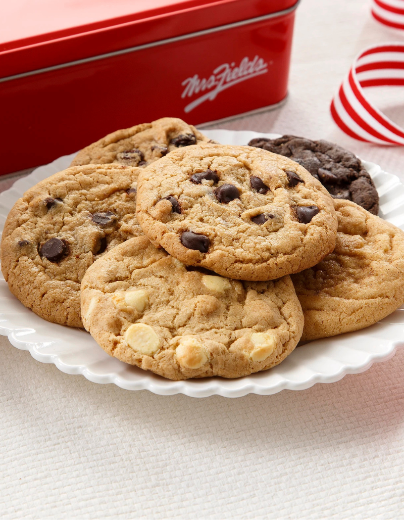 A pile of assorted Mrs. Fields cookies on a white scalloped plate, with a red Mrs.