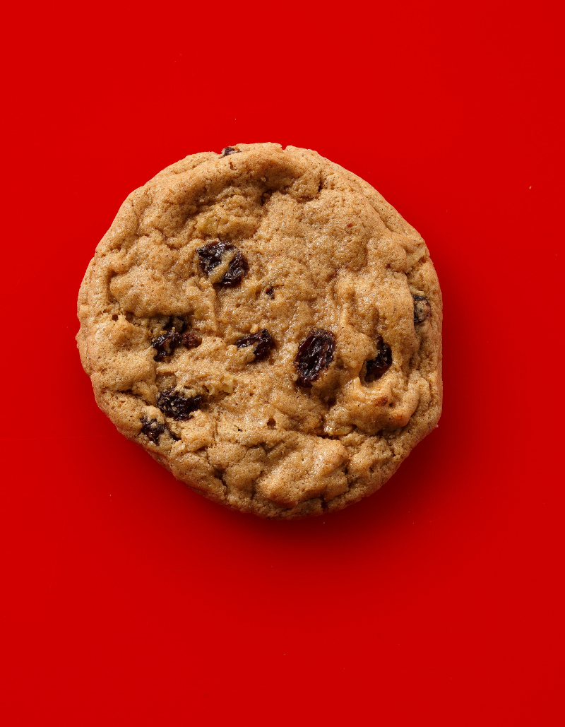 A single round chocolate chip cookie with dark raisin-like pieces, photographed from above on a solid bright red background.