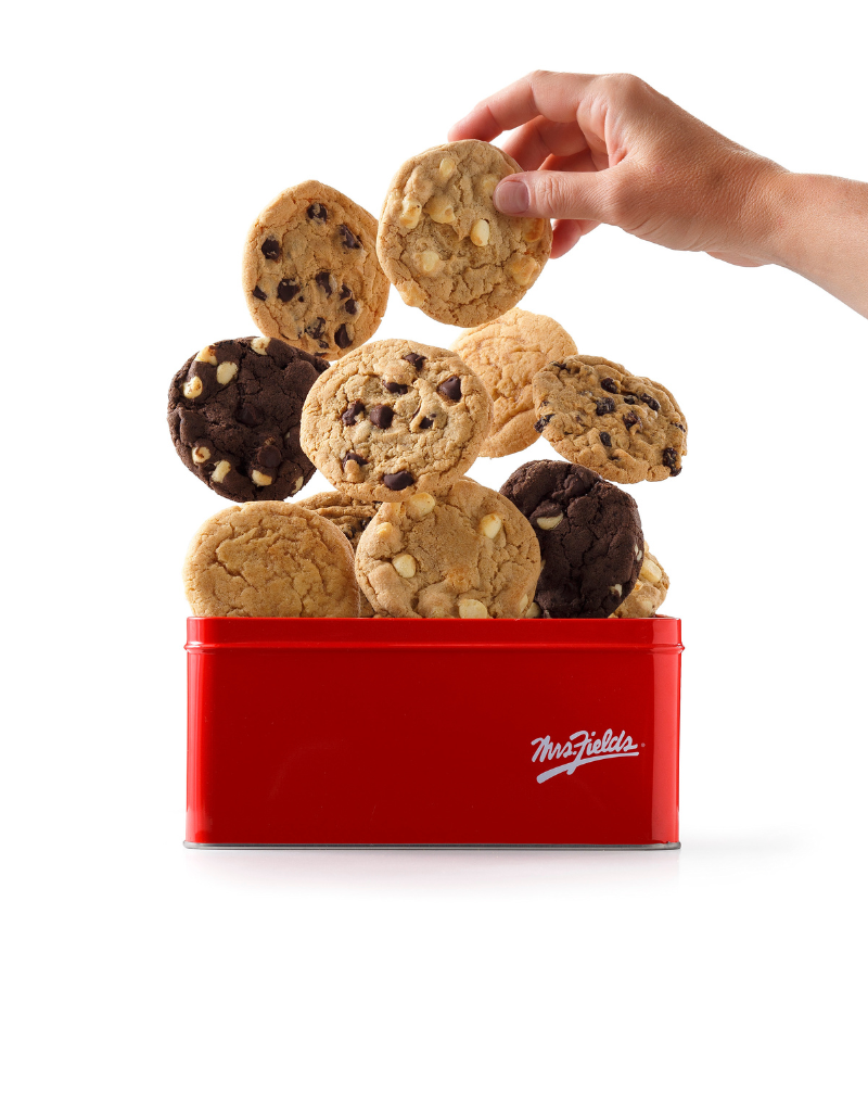 A red Mrs. Fields cookie tin with cookies seemingly jumping out of the top in a variety of flavors. Someone's hand is reaching in to grab one of the floating cookies.