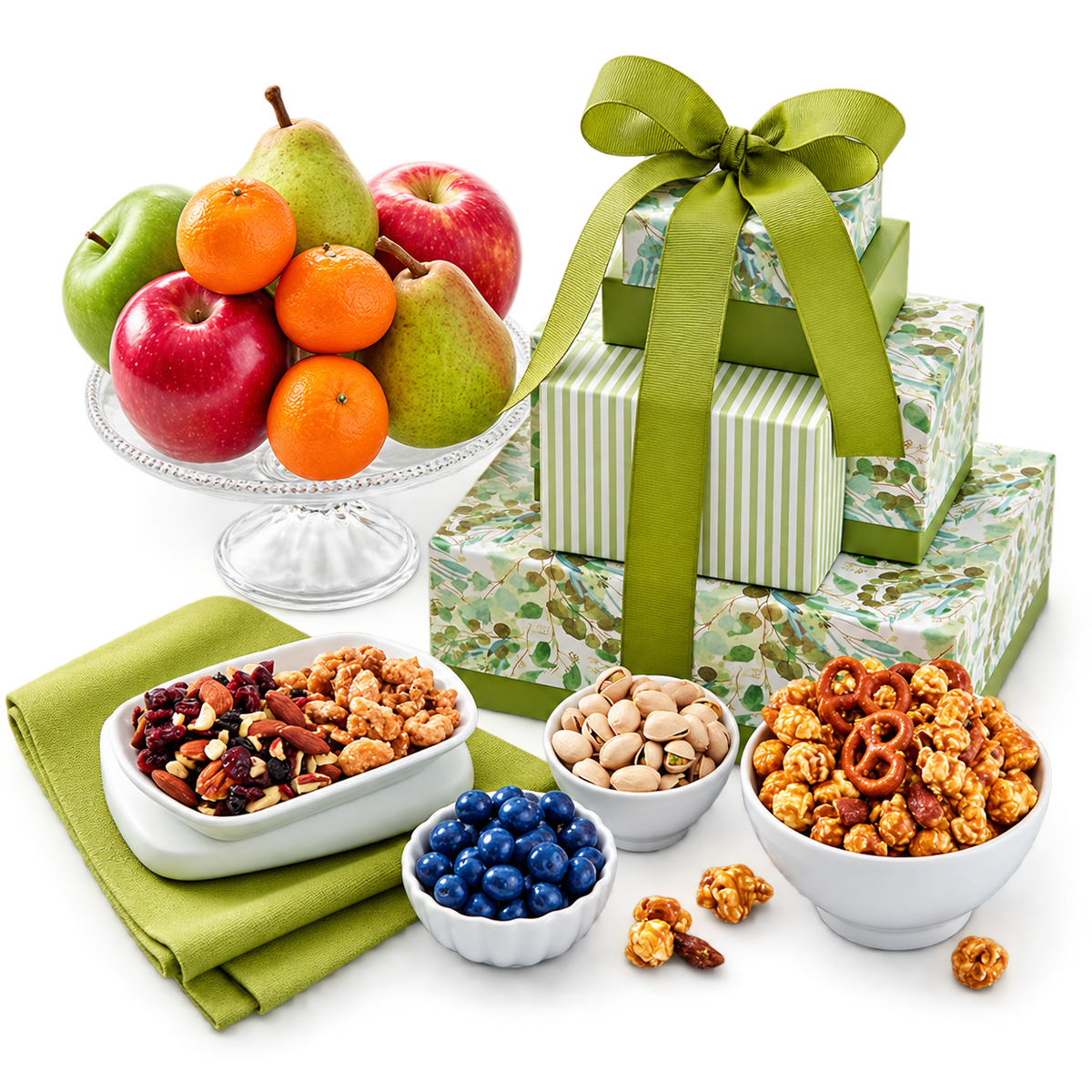 A gift basket arrangement featuring stacked patterned gift boxes tied with a large green ribbon, alongside bowls of snacks and a glass stand of fresh fruit on a white surface.