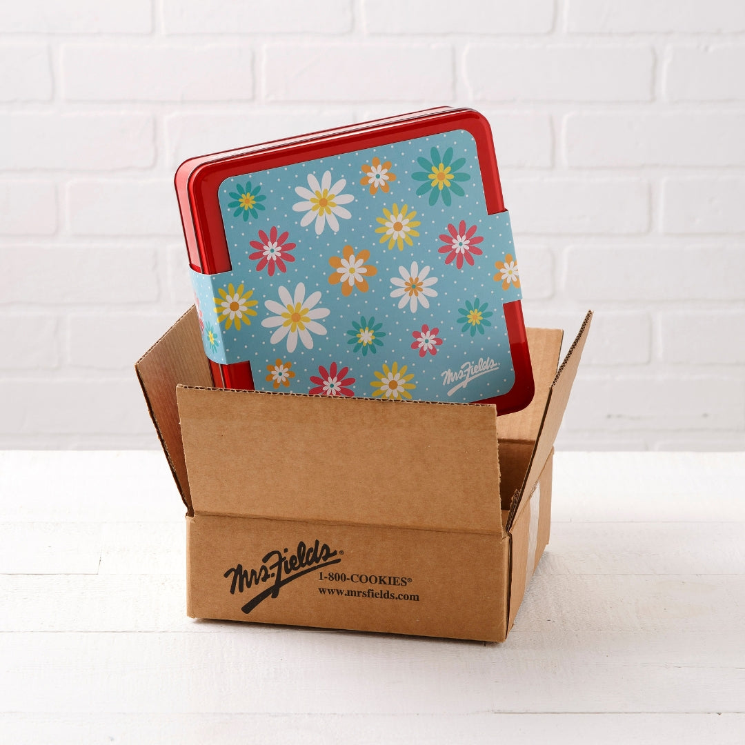 A square red tin with a sleeve on it featuring a colorful pastel daisy pattern partially tucked inside an open brown cardboard mailer box on a white surface against a white brick wall.