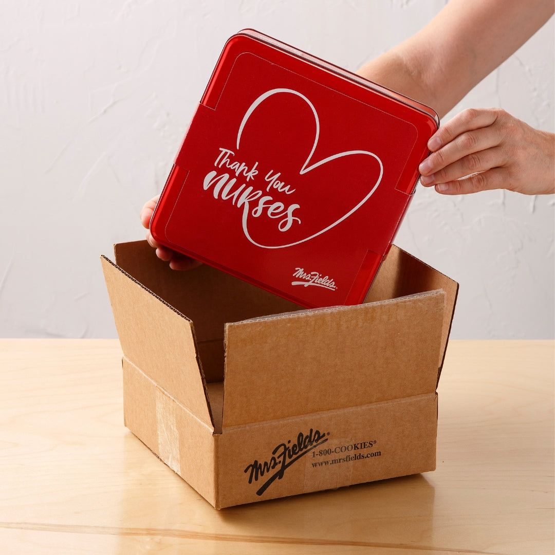 A person's hands are shown holding a red square tin lid above an open brown cardboard box on a light wood table. The lid has a white heart outline and the text 'Thank You Nurses' with a small 'The Gift of Life' logo.