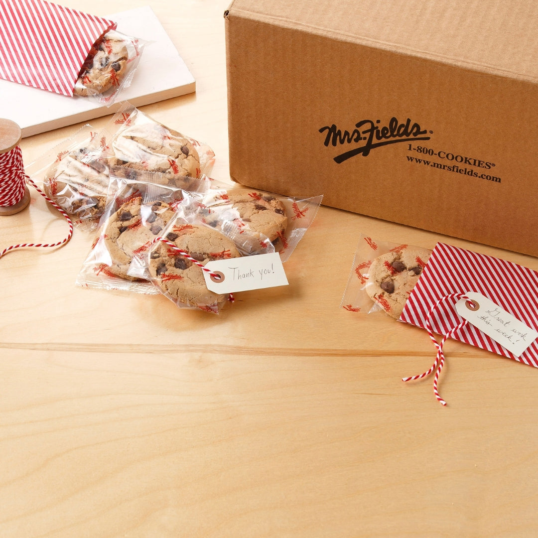 A brown cardboard box printed with the brand name 'Mrs. Fields' and several individually wrapped chocolate chip cookies tied with red-and-white striped twine and holiday tags, arranged on a light wood tabletop.