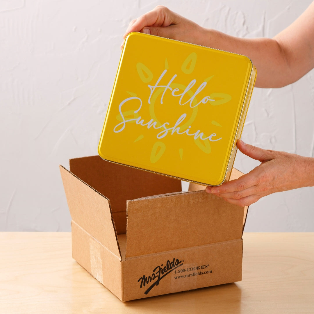 A person holds a square yellow tin lid with the words 'Hello Sunshine' in white script and light green sunburst accents, placing it into an open brown cardboard box on a light wood table against a white wall.