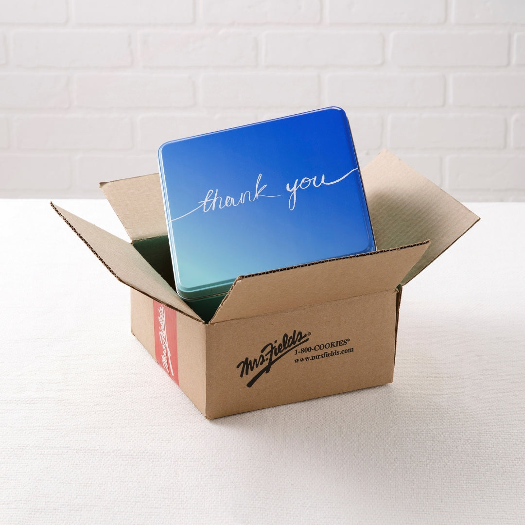 A blue square metal tin with 'thank you' written in white script, partially inside an open brown cardboard box on a white surface against a white brick wall.