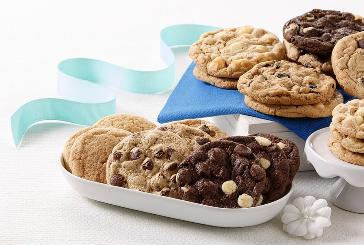 An assortment of chocolate chip cookies in various flavors, including chocolate chip, chocolate white chip, and oatmeal cookie, displayed on white and blue serving platters with decorative ribbons on a white background.