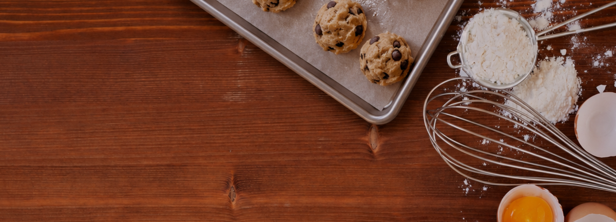raw chocolate chip cookies on a baking pan. flour eggs and a whisk on a wood countertop